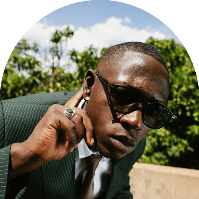 Homme en habit et portant des lunettes soleil, qui tend l'oreille à l'objectif, l'index tendu derrière l'oreille.